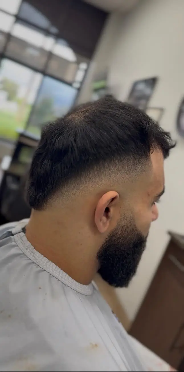 Back-side view of a drop fade curving behind the ear, with a full dark beard